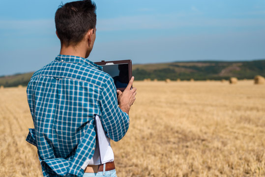 A young agronomist in a white t-shirt and a blue shirt with a tablet and a notebook stands at a haystack, taking pictures of the field