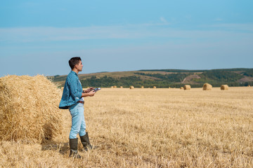 Agronomist farmer in jeans, shirt with a tablet is in the field with a stack, with tablet looking into the distance. Rural business, agricultural industry, freedom after work, concept
