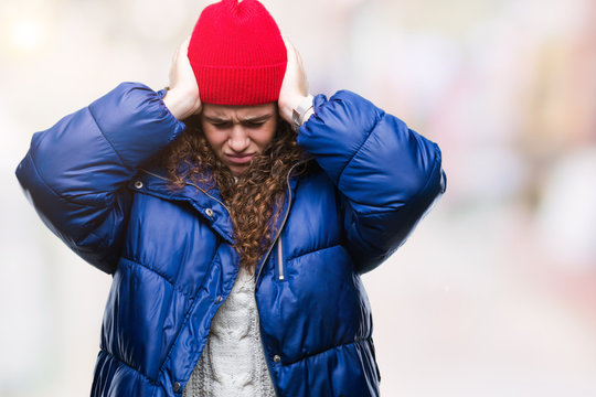 Beautiful young brunette curly hair girl wearing winter coat, wool cap and sweater over isolated background suffering from headache desperate and stressed because pain and migraine. Hands on head.