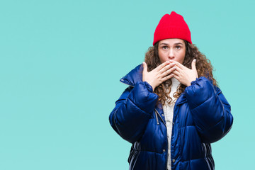 Beautiful young brunette curly hair girl wearing winter coat, wool cap and sweater over isolated background shocked covering mouth with hands for mistake. Secret concept.