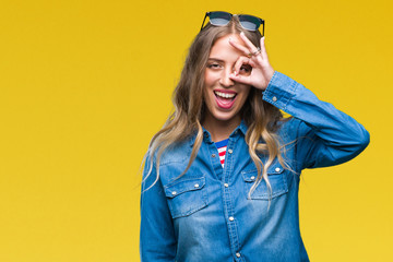 Beautiful young blonde woman wearing sunglasses over isolated background doing ok gesture with hand smiling, eye looking through fingers with happy face.
