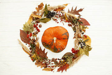 Autumn Flat Lay. Happy Thanksgiving. Pumpkin in beautiful fall leaves wreath with berries,nuts,acorns, herbs on rustic white background top view. Seasons greetings card mockup