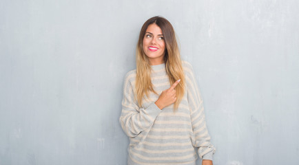 Fototapeta na wymiar Young adult woman over grunge grey wall wearing winter sweater with a big smile on face, pointing with hand and finger to the side looking at the camera.