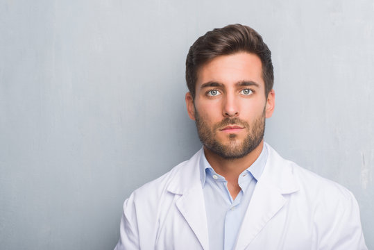 Handsome young professional man over grey grunge wall wearing white coat with serious expression on face. Simple and natural looking at the camera.