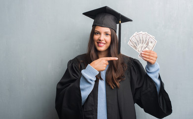 Young brunette woman over grunge grey wall wearing graduate uniform holding dollars very happy...