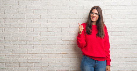 Fototapeta na wymiar Young brunette woman standing over white brick wall doing happy thumbs up gesture with hand. Approving expression looking at the camera with showing success.