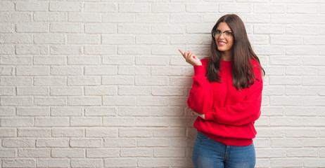 Young brunette woman standing over white brick wall with a big smile on face, pointing with hand and finger to the side looking at the camera.