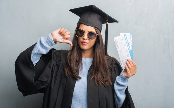 Young brunette woman over grunge grey wall wearing graduate uniform holding boarding pass with angry face, negative sign showing dislike with thumbs down, rejection concept