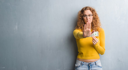 Young redhead woman over grey grunge wall holding a dollar with open hand doing stop sign with...