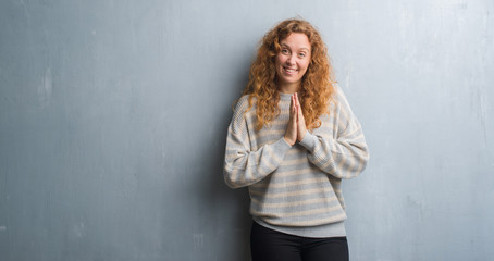 Fototapeta premium Young redhead woman over grey grunge wall praying with hands together asking for forgiveness smiling confident.