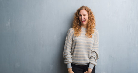 Young redhead woman over grey grunge wall winking looking at the camera with sexy expression, cheerful and happy face.