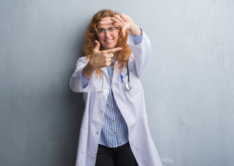 Young redhead doctor woman over grey grunge wall wearing a coat smiling making frame with hands and fingers with happy face. Creativity and photography concept.