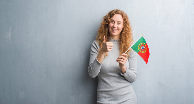 Young redhead woman over grey grunge wall holding flag of Portugal happy with big smile doing ok sign, thumb up with fingers, excellent sign