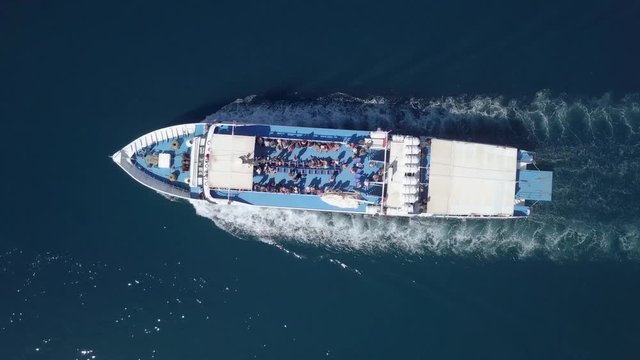 Following a medium sized Ferry as it is approaching a greek island, with people on the upper deck - Top down aerial footage.