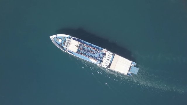 Following a medium sized Ferry as it is approaching a greek island, with people on the upper deck - Top down aerial footage.