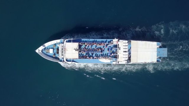 Following a medium sized Ferry as it is approaching a greek island, with people on the upper deck - Top down aerial footage.