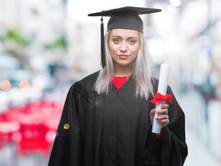 Young blonde woman wearing graduate uniform holding degree over isolated background with a confident expression on smart face thinking serious