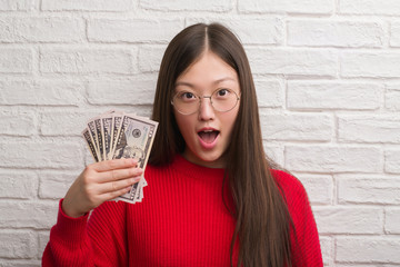 Young Chinese woman over brick wall holding dollars scared in shock with a surprise face, afraid and excited with fear expression