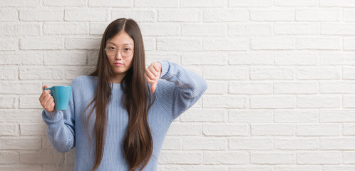 Young Chinise woman drinking a cup of coffee over white brick wall with angry face, negative sign showing dislike with thumbs down, rejection concept