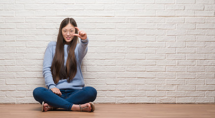 Young Chinese woman sitting on the floor over brick wall smiling and confident gesturing with hand doing size sign with fingers while looking and the camera. Measure concept.