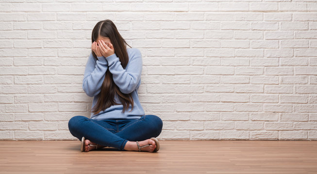 Young Chinese woman sitting on the floor over brick wall with sad expression covering face with hands while crying. Depression concept.