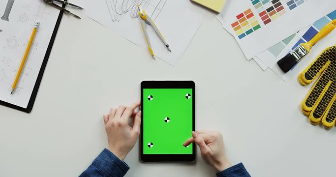 View from above on the female hands scrolling and tapping on the chroma key screen of the black vertical tablet device on the designer desk with drawings stuff. Green screen. Tracking motion.