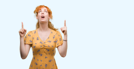 Young redhead woman amazed and surprised looking up and pointing with fingers and raised arms.
