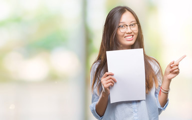Young asian woman holding blank paper over isolated background very happy pointing with hand and finger to the side