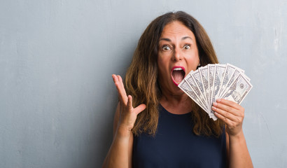 Middle age hispanic woman standing over grey grunge wall holding dollars very happy and excited, winner expression celebrating victory screaming with big smile and raised hands