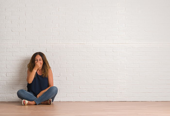 Middle age hispanic woman sitting on the floor over white brick wall smelling something stinky and disgusting, intolerable smell, holding breath with fingers on nose. Bad smells concept.