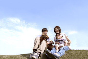 青空バックにペットの犬を抱き芝生に座る幸せな家族。家族、親子、幸せ、愛情、育児、ペットイメージ