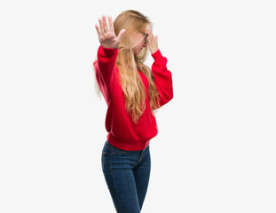 Obraz na płótnie Canvas Blonde teenager woman wearing red sweater covering eyes with hands and doing stop gesture with sad and fear expression. Embarrassed and negative concept.