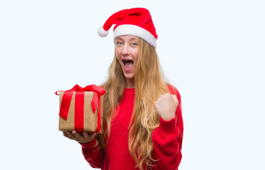 Blonde teenager woman wearing santa claus hat screaming proud and celebrating victory and success very excited, cheering emotion