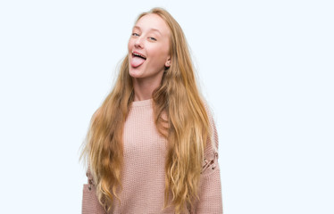 Blonde teenager woman wearing pink sweater sticking tongue out happy with funny expression. Emotion concept.