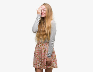 Blonde teenager woman wearing flowers skirt covering one eye with hand with confident smile on face and surprise emotion.