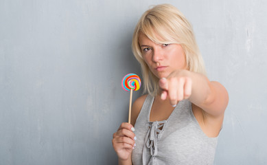 Caucasian adult woman over grey grunge wall eating candy lollipop pointing with finger to the camera and to you, hand sign, positive and confident gesture from the front