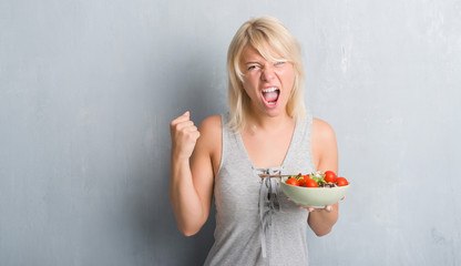 Adult caucasian woman over grunge grey wall eating tomato salad annoyed and frustrated shouting with anger, crazy and yelling with raised hand, anger concept