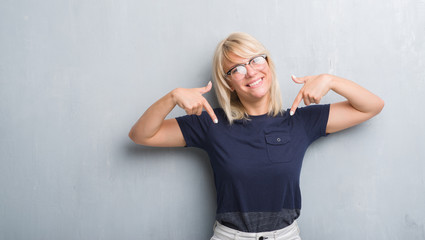 Adult caucasian woman over grunge grey wall wearing glasses looking confident with smile on face, pointing oneself with fingers proud and happy.