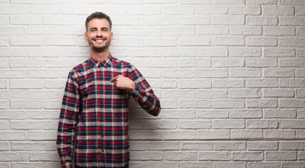 Young adult man over white brick wall with surprise face pointing finger to himself