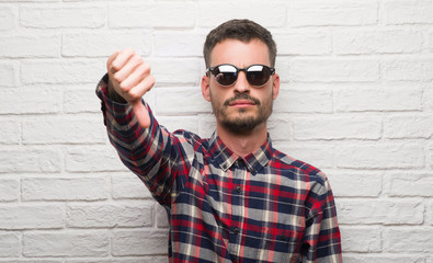 Young adult man wearing sunglasses standing over white brick wall with angry face, negative sign...