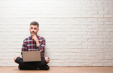 Young adult man over brick wall using laptop serious face thinking about question, very confused...