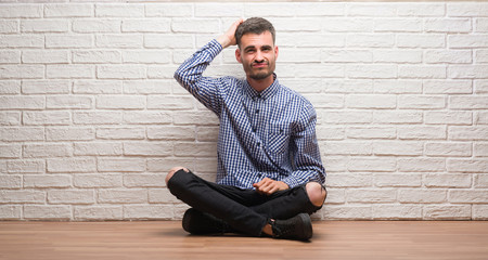 Young adult man sitting over white brick wall confuse and wonder about question. Uncertain with doubt, thinking with hand on head. Pensive concept.