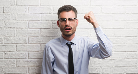 Young adult business man standing over white brick wall annoyed and frustrated shouting with anger,...