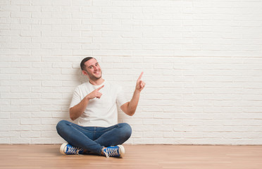 Young caucasian man sitting on the floor over white brick wall smiling and looking at the camera...