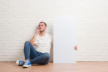Young caucasian man sitting over white brick wall holding bottle of water serious face thinking...