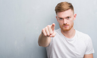 Young redhead man over grey grunge wall pointing with finger to the camera and to you, hand sign, positive and confident gesture from the front