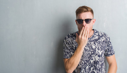 Young redhead man over grey grunge wall wearing sunglasses cover mouth with hand shocked with shame for mistake, expression of fear, scared in silence, secret concept