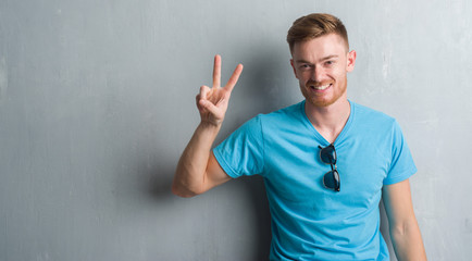 Fototapeta premium Young redhead man over grey grunge wall wearing casual outfit showing and pointing up with fingers number two while smiling confident and happy.