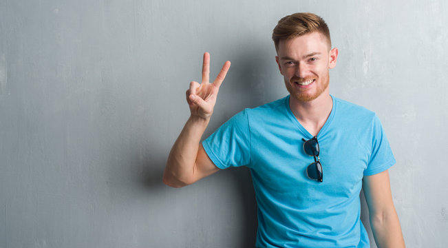 Young redhead man over grey grunge wall wearing casual outfit showing and pointing up with fingers number two while smiling confident and happy.