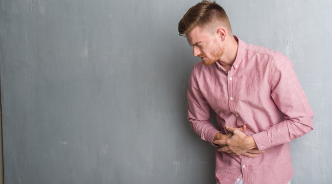 Young redhead man over grey grunge wall wearing pink shirt with hand on stomach because indigestion, painful illness feeling unwell. Ache concept.